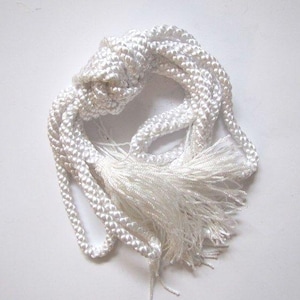 Puede incluir: Un cordón decorativo blanco con un centro anudado y una borla en un extremo. El cordón está hecho de un material grueso y trenzado, probablemente para decoración o como accesorio. La borla está hecha de muchos hilos finos del mismo material blanco.