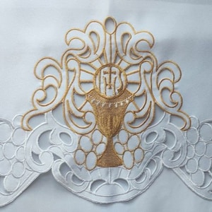 Puede incluir: Tela blanca con bordado dorado de un cáliz con una cruz y un diseño de sol. La tela tiene un borde festoneado con un patrón floral.