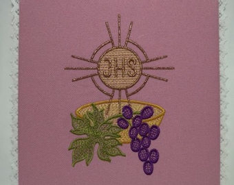May include: Pink fabric with white lace trim, featuring an embroidered design of a chalice, grapes, and a sunburst with the letters "JHS" in the center.