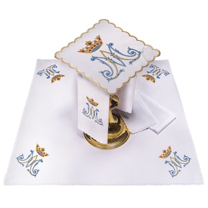Può includere: Biancheria da altare bianca con una "M" ricamata in blu e oro con una corona. Le lenzuola sono disposte su un tavolo con un calice d'oro.