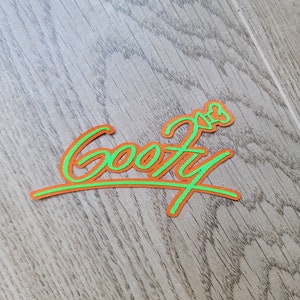 May include: A green and orange embroidered patch with the word "Goofy" in a cursive font.