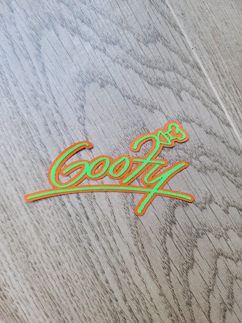 May include: A green and orange embroidered patch with the word "Goofy" in a cursive font.