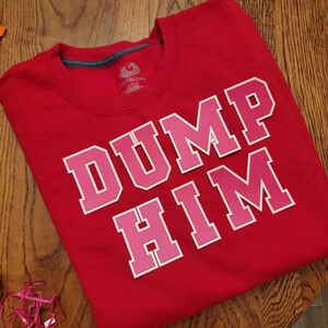 Peut inclure: T-shirt rouge avec les mots "DUMP HIM" en lettres roses avec un contour blanc.