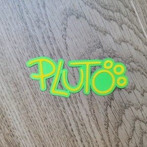 May include: A green and yellow felt cutout of the word "Pluto" with a yellow outline.
