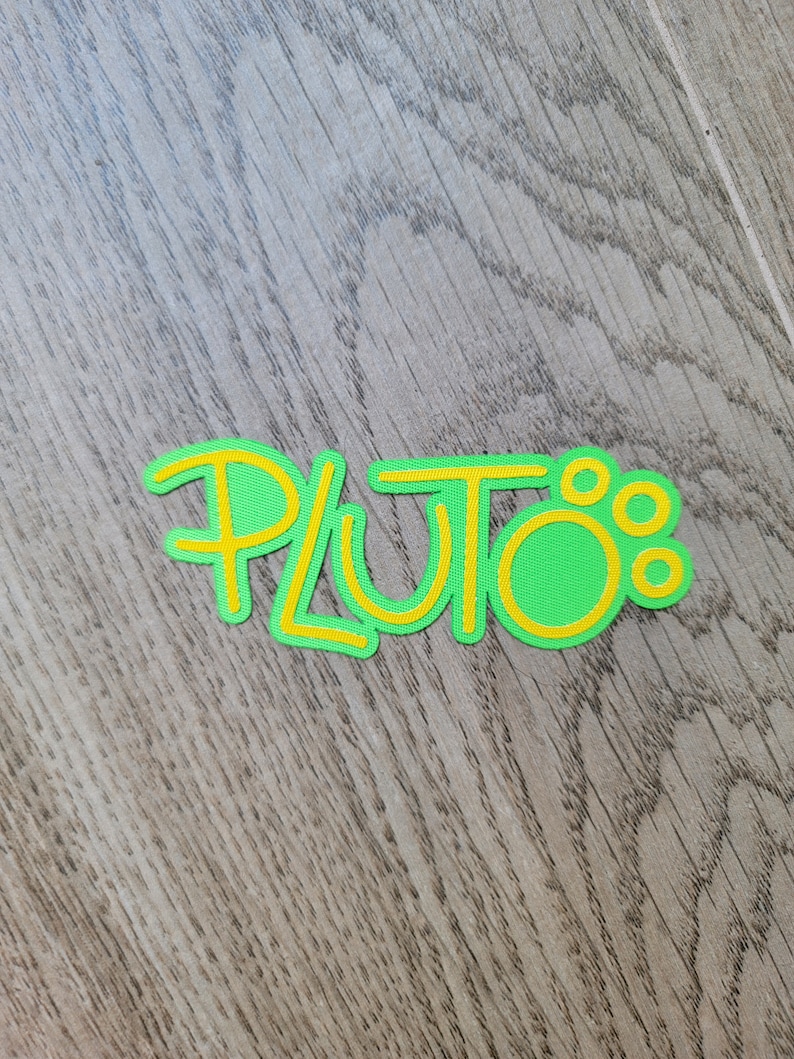 May include: A green and yellow felt cutout of the word "Pluto" with a yellow outline.