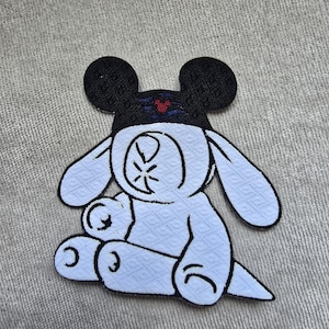 May include: A white embroidered patch featuring a cartoon character with a Mickey Mouse-style hat. The hat is black with a red and blue design. The character has large ears and is sitting down.