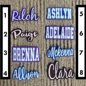 Peut inclure: Huit étiquettes de nom personnalisées avec des lettres blanches sur des fonds de couleurs variées. Les noms sont : Rilah, Paige, Brenna, Allison, Ashlyn, Adelaide, Mckenna et Clara.