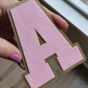 May include: A pink felt letter A with a brown outline. The letter is held in a hand.