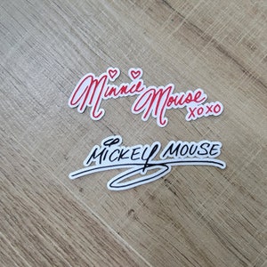 Pode incluir: Dois adesivos de assinatura vermelhos e pretos com os nomes "Minnie Mouse xoxo" e "Mickey Mouse" em itálico.