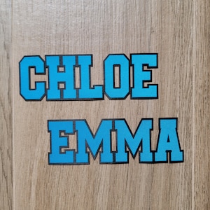 May include: Two names, "CHLOE" and "EMMA", are spelled out in large, blue, block letters with black outlines. The letters are on a wood-grain background.