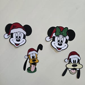 Op de afbeelding: Vier cartoon karakter uitsparingen: Mickey Mouse, Minnie Mouse, Pluto en Goofy, allemaal met kerstmutsen. Mickey en Minnie hebben zwarte oren en witte gezichten. Pluto is geel en Goofy is bruin. De uitsparingen staan op een witte achtergrond.