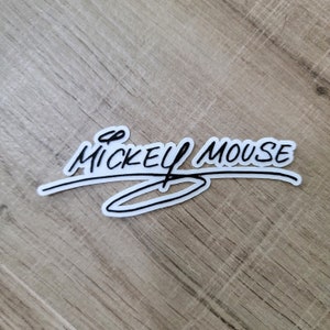 May include: White sticker with black text that reads "Mickey Mouse" in a handwritten style. The text is curved and has a circle underneath it.