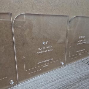 Peut inclure: Trois gabarits de rayon d'angle en acrylique transparent avec des bords arrondis. Chaque gabarit comporte du texte gravé incluant R 1", R 1-1/2", Ready Laser, et des instructions pour aligner avec le bord et le centre de la planche.