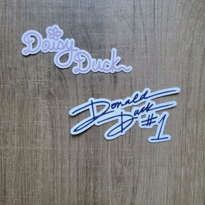 May include: Two stickers on a wood background. The top sticker is pink and says "Daisy Duck". The bottom sticker is blue and says "Donald Duck #1".