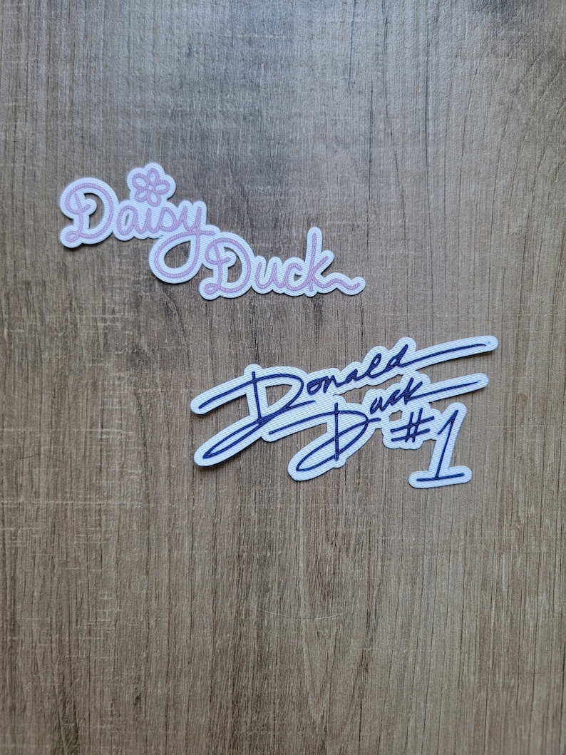 May include: Two stickers on a wood background. The top sticker is pink and says "Daisy Duck". The bottom sticker is blue and says "Donald Duck #1".