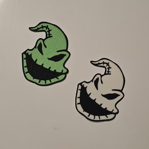 May include: Two patches featuring a stylized face with a stitched design. One patch is green with a black outline and black mouth. The other patch is white with a black outline and black mouth. The patches are on a white background.