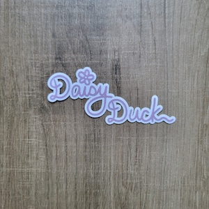 May include: A white sticker with the text "Daisy Duck" in pink cursive lettering. A small pink flower is above the word "Daisy".