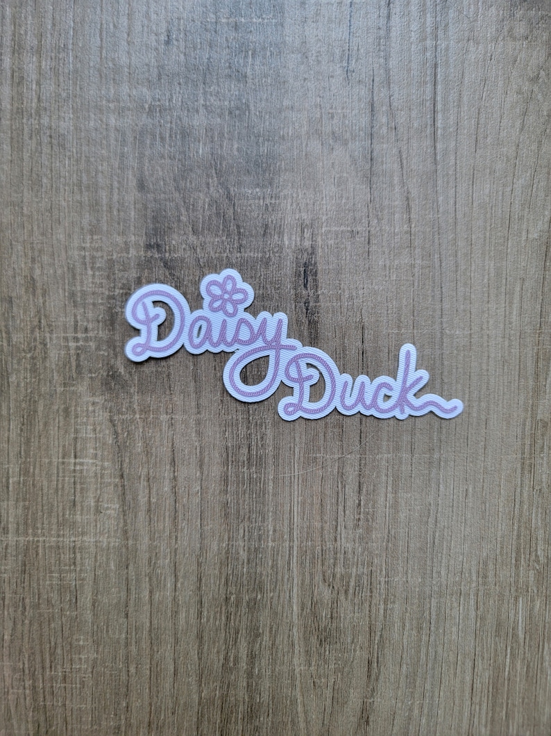 May include: A white sticker with the text "Daisy Duck" in pink cursive lettering. A small pink flower is above the word "Daisy".