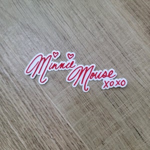 May include: White sticker with red lettering that says "Minnie Mouse xoxo" with two red hearts above the name.