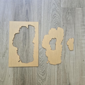 May include: Three wooden cutout shapes of different sizes, all in the shape of a lake. The largest cutout is on the left, the medium cutout is on the right, and the smallest cutout is to the right of the medium cutout. All three cutouts are on a light gray wood floor.
