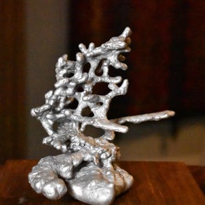 May include: A silver-colored metallic sculpture with an organic, branching structure resembling coral or tree roots. The object features a textured, uneven surface with multiple interconnected protrusions and open spaces throughout its form.