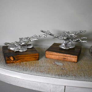 May include: Two silver-colored metal sculptures shaped like branching coral or twigs, each mounted on a square, dark brown wooden base. The metallic structures have a textured, organic appearance and are connected by a thin, horizontal metal branch.