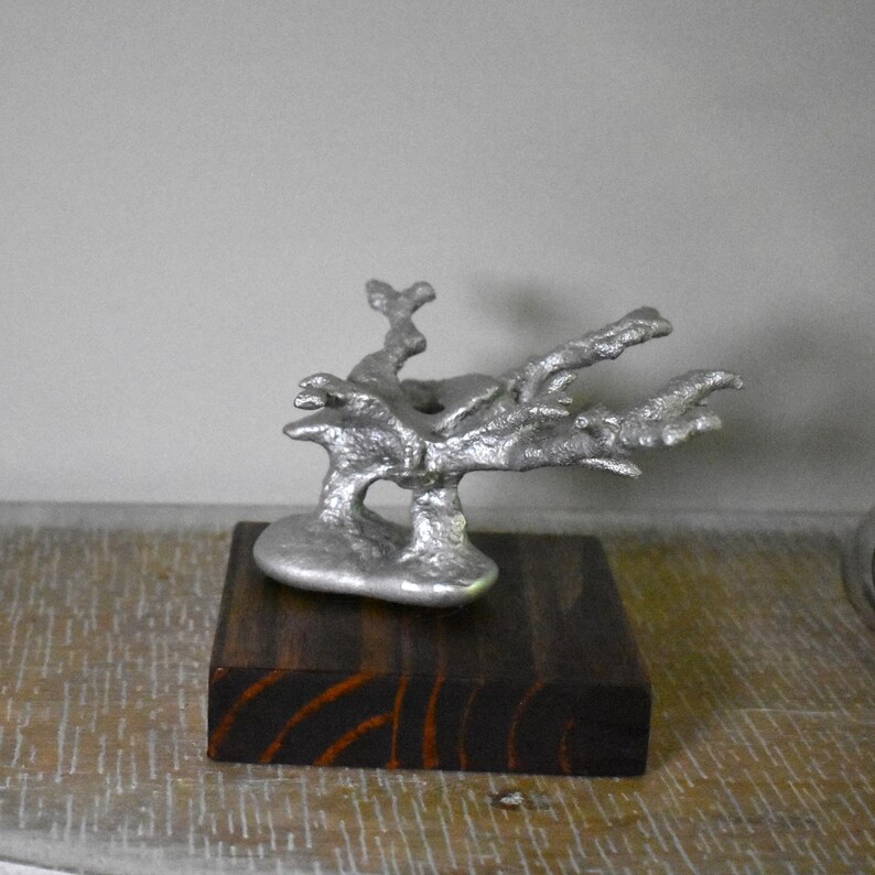 May include: A silver-colored, textured metal sculpture shaped like a branching tree or coral formation, mounted securely onto a square, dark brown wooden base with visible wood grain patterns throughout the block's surface.