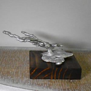May include: A silver-colored metal sculpture with a textured, branch-like form extending horizontally from a flat, irregular base. The metal piece is securely mounted onto a square, dark brown wooden block with visible wood grain patterns.