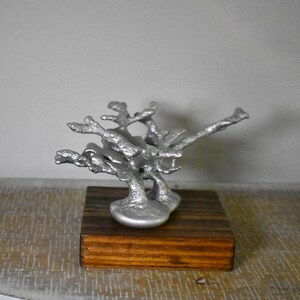 May include: A silver-colored, textured, branch-like metal sculpture mounted on a square, dark brown wooden base. The sculpture features multiple irregular, protruding arms that extend outward from a central, solid, rounded metal foundation.