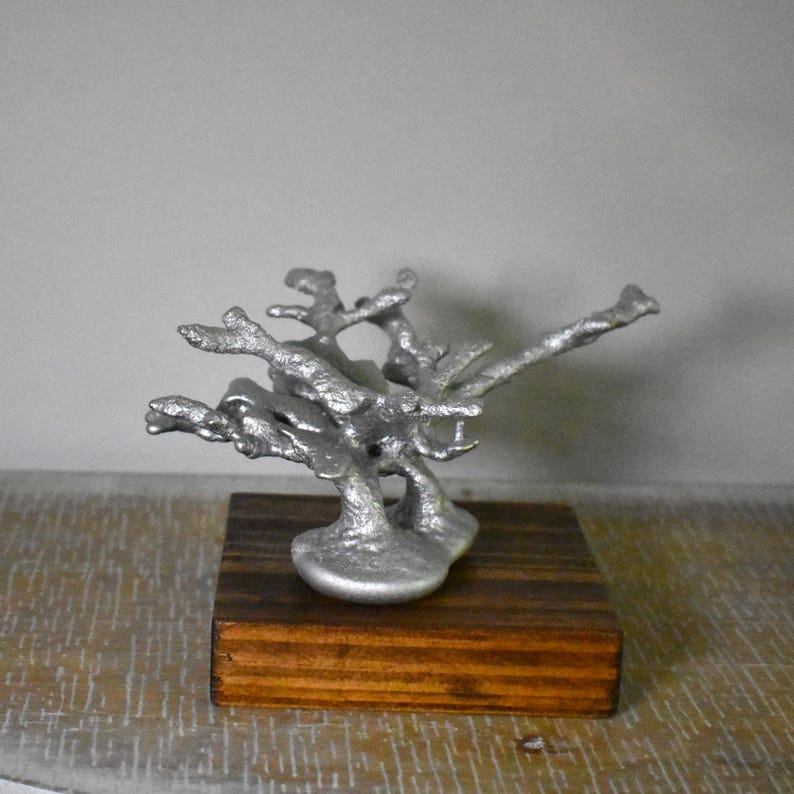 May include: A silver-colored, textured, branch-like metal sculpture mounted on a square, dark brown wooden base. The sculpture features multiple irregular, protruding arms that extend outward from a central, solid, rounded metal foundation.
