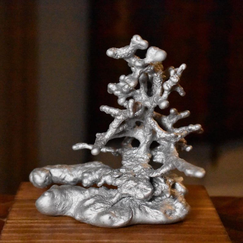 May include: A silver-colored metallic sculpture with a textured, organic, and branching form that resembles a small tree or coral structure. The object has a molten, uneven surface with rounded protrusions and a solid, wide base.