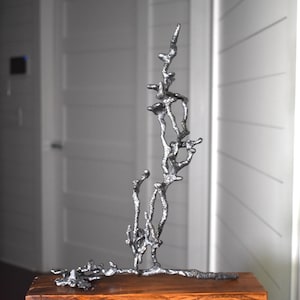 May include: A silver metal sculpture of a tree with a unique, organic, and abstract design. The sculpture is displayed on a wooden base.