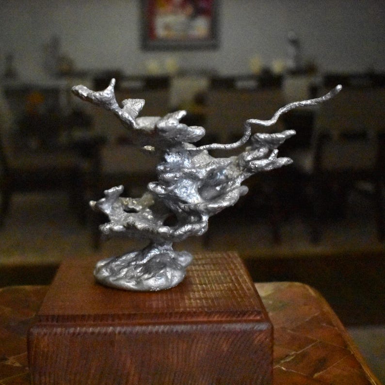 May include: A silver-colored, abstract metal sculpture with a porous, organic, and branching structure. The sculpture is mounted on a square, dark brown wooden base, featuring a textured surface with irregular protrusions and a metallic sheen.