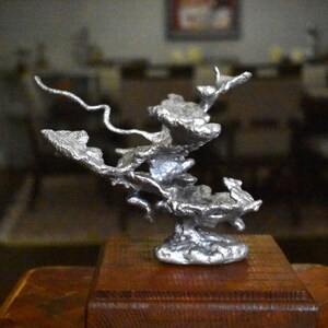 May include: A silver-colored, abstract metal sculpture with a jagged, organic, and porous texture. The sculpture features multiple branching, wing-like protrusions extending from a central base, mounted on a square, dark brown wooden block.