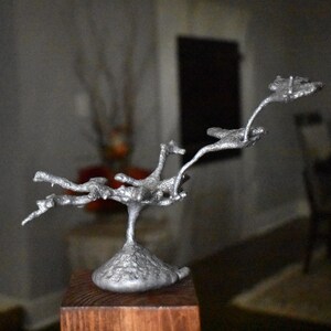 May include: A silver-colored metal sculpture featuring a textured, organic branch-like structure extending from a solid, conical base. The object has a rough, cast-metal finish with multiple protruding arms that reach upward and outward in a dynamic form.