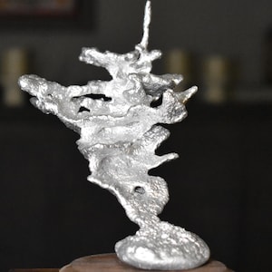 May include: A silver-colored, abstract sculpture with a textured surface. The sculpture has an organic, branching form, mounted on a small, round, wooden base. The artwork has a unique, artistic design.