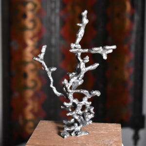 May include: A silver-colored, abstract sculpture resembling a tree or coral. The sculpture is mounted on a small, rectangular wooden base. The background features a blurred pattern of red, orange, and black.