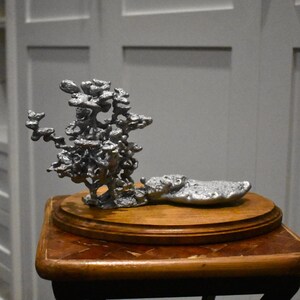 May include: A silver-colored metal sculpture with an organic, branching, and porous structure that resembles a tree or coral formation, attached to a flat, textured base, all mounted on a smooth, oval-shaped wooden display stand.