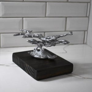 May include: A silver-colored, organic-shaped metal sculpture with a textured, branch-like appearance mounted on a rectangular, dark-stained wooden base. The metallic structure features multiple protruding, irregular arms extending horizontally from a central point.