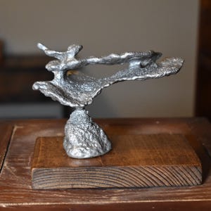 May include: A silver-colored abstract sculpture with a textured surface, mounted on a rectangular wooden base. The sculpture has an organic, flowing shape, resembling a cloud or a stylized form. The wooden base is a dark brown color.