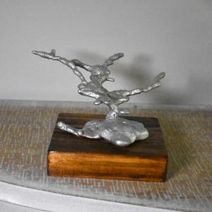 May include: A silver-colored metal sculpture with an irregular, organic, branch-like form mounted on a square, dark brown wooden base. The metal piece features a textured, molten appearance with multiple protruding arms extending outward.