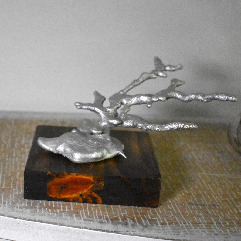 May include: A silver-colored metal sculpture shaped like a branching coral structure, mounted on a square, dark brown wooden base. The metal has a textured, organic surface, and the wooden base features a distinct, lighter orange-brown grain pattern.