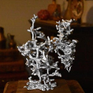 May include: A silver-colored metal sculpture featuring an intricate, branching, organic structure with numerous small, rounded protrusions and interconnected segments, mounted on a rectangular wooden base, creating a complex, porous, and textured form.