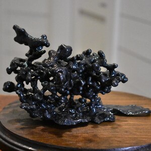 May include: A dark, metallic sculpture with a porous, organic, and irregular structure featuring numerous interconnected nodules and branches. The object has a glossy, reflective finish and is mounted on a circular, dark brown wooden base.
