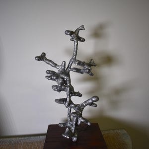 May include: A silver-colored metal sculpture with an abstract, branching design. The sculpture is mounted on a dark brown wooden base. The artwork has a textured surface and is displayed against a neutral background.