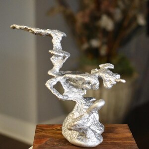 May include: A silver-colored abstract sculpture with an organic, flowing design. The sculpture is mounted on a rectangular wooden base. The artwork has a textured surface and an irregular shape, suggesting a natural form.