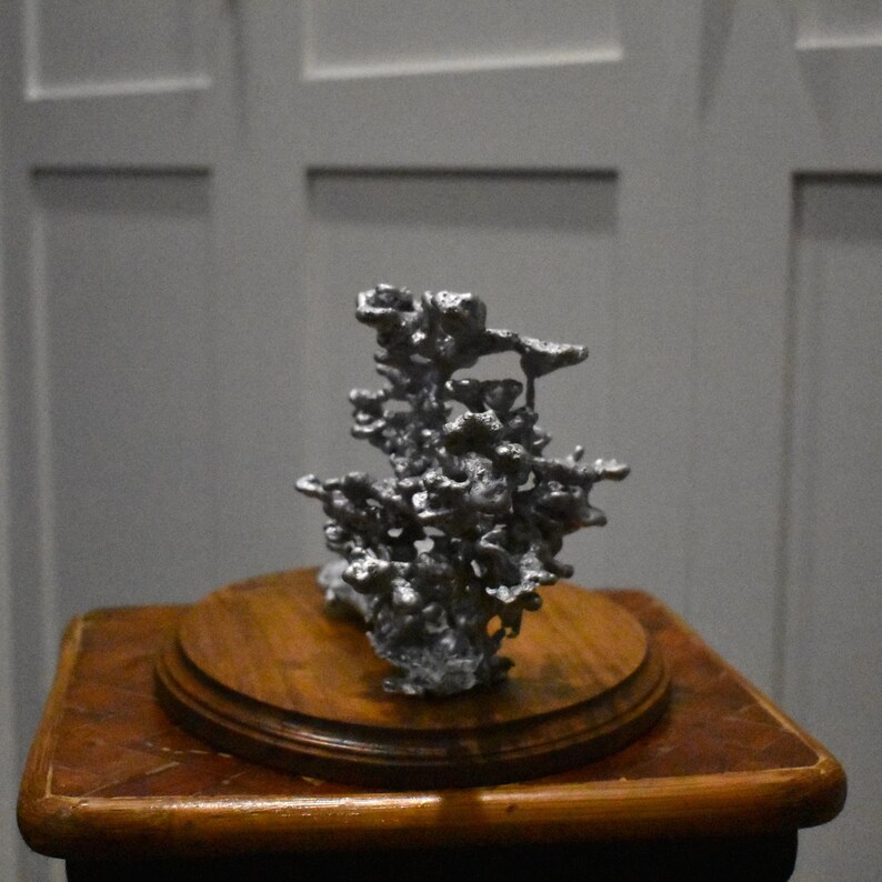 May include: A silver-colored, irregular, porous metal sculpture with a rough, textured surface. The sculpture is mounted on a circular, polished wooden base that features a warm, medium-brown tone and a smooth, rounded edge finish.