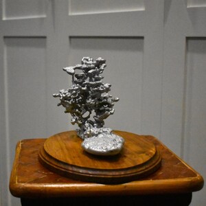 May include: A silver-colored metal sculpture with an irregular, porous, and organic structure resembling a tree or coral formation, mounted on a circular, polished wooden base with a warm brown tone and visible wood grain patterns.