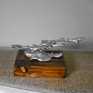 May include: A silver-colored metal sculpture with an irregular, organic, branch-like form mounted on a rectangular, dark brown wooden base. The metal piece features a textured, molten appearance with multiple protruding arms extending horizontally.