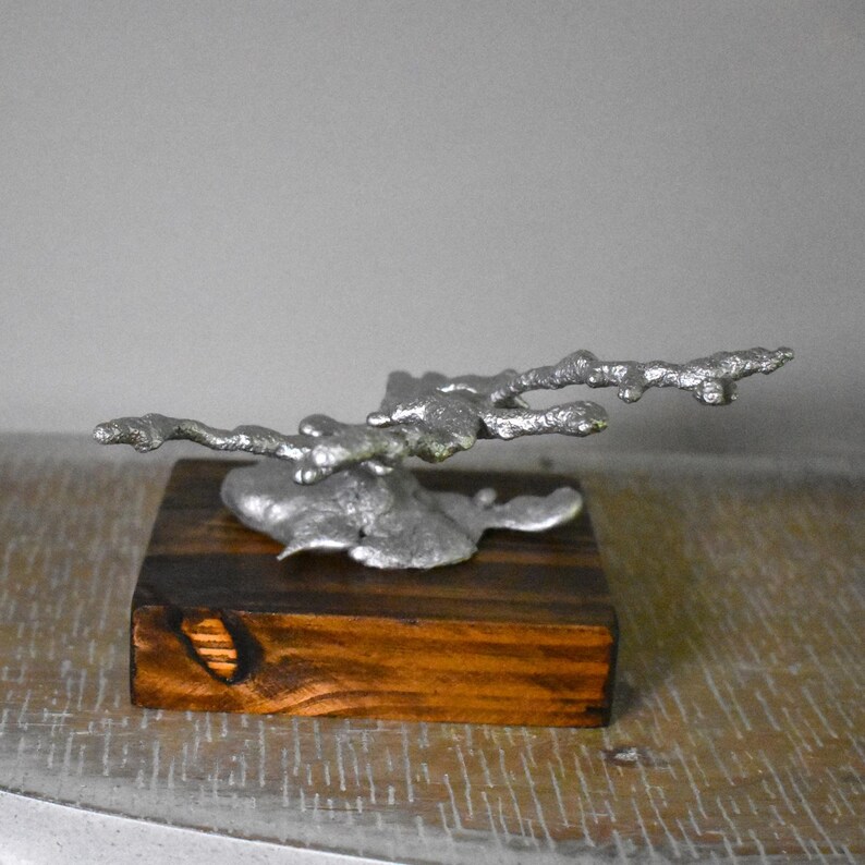 May include: A silver-colored metal sculpture with an irregular, organic, branch-like form mounted on a rectangular, dark brown wooden base. The metal piece features a textured, molten appearance with multiple protruding arms extending horizontally.